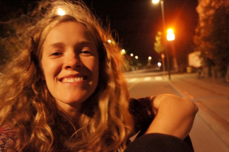 foto. smilende kvinne i helfigur fotografert i halvmørke i en gate med gatelys i bakgrunnen