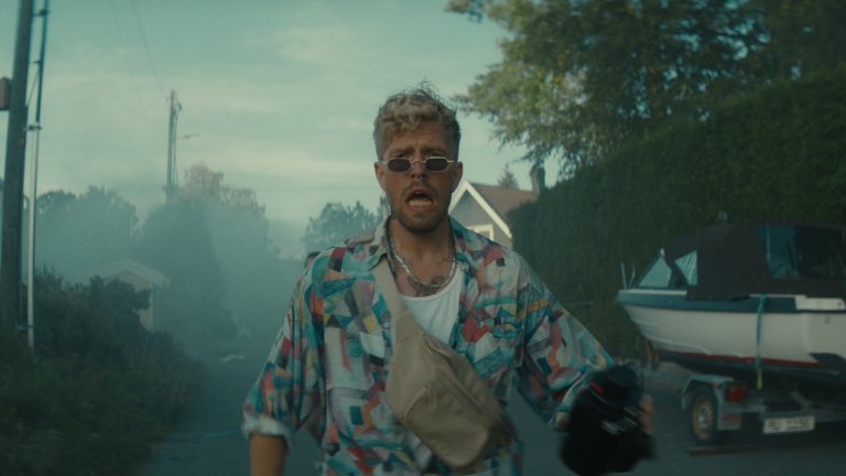 foto. mann med små solbriller og mønstrete skjorte går nedover en villavei mot kamera mens han roper