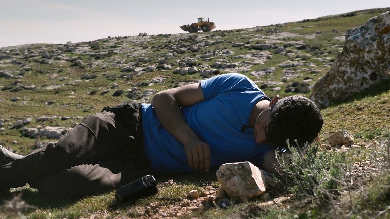 foto. en mann i tskjorte ligger på en gressbakke med en gravemaskin i bakgrunnen