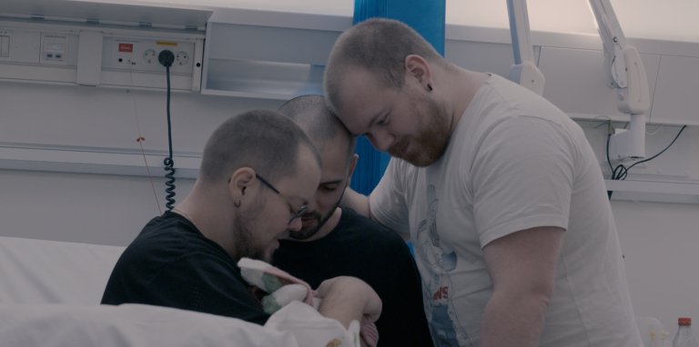foto. tre menn med kort hår og skjekk er samla rundt ein baby ved ei sjukehusseng