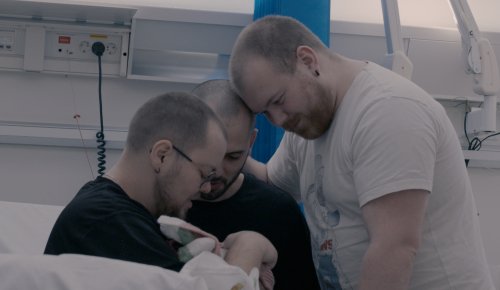 foto. tre menn med kort hår og skjekk er samla rundt ein baby ved ei sjukehusseng