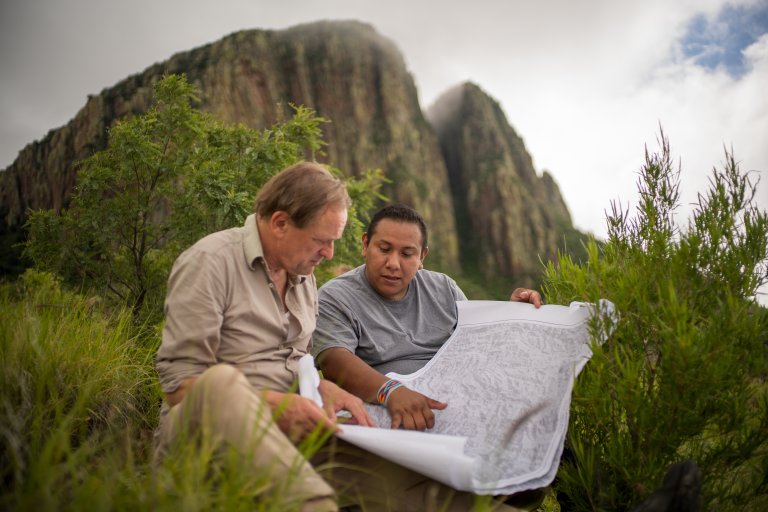 foto. to menn sitter i grønne busker foran et fjell og studerer et kart