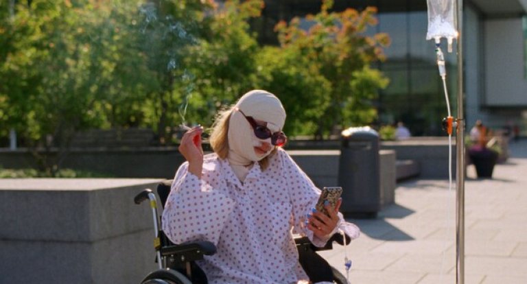 Bilde fra Syk pike. Kvinne sitter med bandasje over hele hodet og røyker i en rullestol mens hun får drypp.