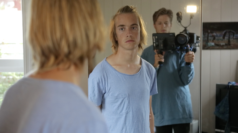 Foto: Bilde fra dokumentaren Nattebarn. Vi ser en gutt med halvlangt blondt hår som står og ser seg i speilet. Han ser alvorlig ut .I bakgrunnen ser vi en mann stå og filme.