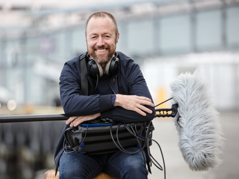 Foto: Bilde av en mann, regissør Håvard Bustnes. Han sitter med lydutstyr og en stor mikrofon-boom på fanget. Han har helt kort hår og kort skjegg. Han er iført olabukse og en blå jakke.