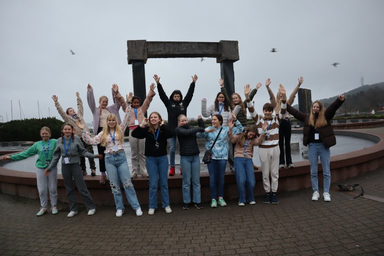 Foto: 16 barn er avbildet ved fonteneskulpturen laget av Kjell Nupen i Otterdalsparken i Kristiansand. Ni ban står på bakken foran skulpturen. Flere av dem strekker armene i været. Syv barn står oppe på fontenekanten og strekker armene i været. I luften ser vi fire måker sikle over fontene.