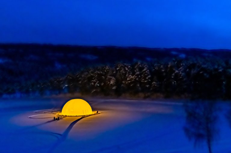 Foto: Bildet viser en islagt fjord i skumring. Man kan skimte snødekt skog og fjell i bakgrunnen. Isen ser nærmest blå ut i mørket og er dekket av snø. Man ser en stor globuslignende kule på isen som er lyst opp innenfra og lyser gult. Et par mennesker kan også skimtes utenfor globen.