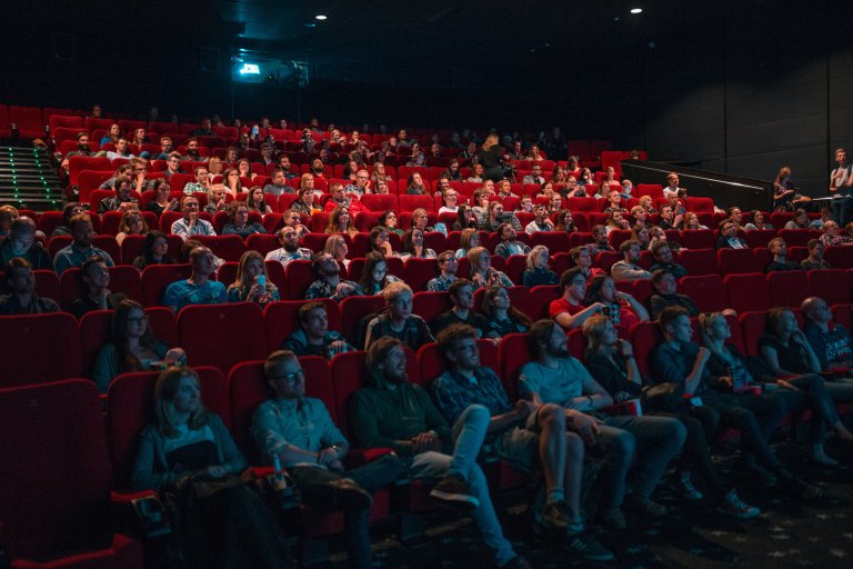 Illustrasjonsbilde: Bildet viser en kinosal med røde seter, ganske full av mannesker. Lyst er delvis på og man ser lyset fra maskinrommet bakerst i salen.