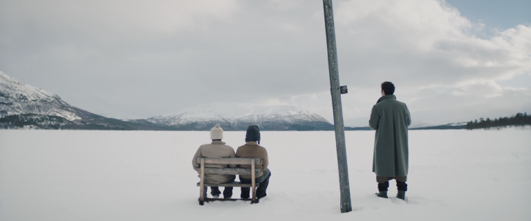 foto. to personer sitter med ryggen til kamera på en benk i et snødekt landskap. til høyre ved en lyktestolpe står en tredje person med ryggen til