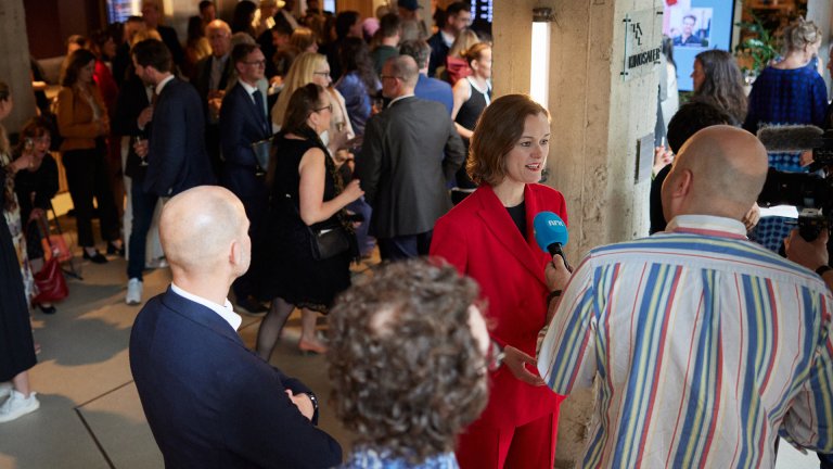 foto. kvinne i rød buksedress midt i bildet blir intervjuet av mann med mikrofon med NRK logo og er omgitt av publikum i dress og kjoler