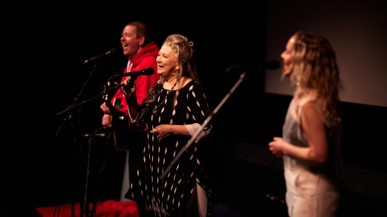 Foto. tre personer står og synger på scenen en mann med gitar til venstre og to kvinner til høyre for ham
