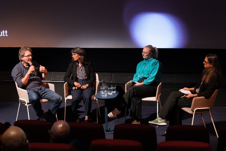 foto. fire personer sitter i hver sin stol på scene, en mann til venstre og tre kvinner, mann snakker i mikrofonen