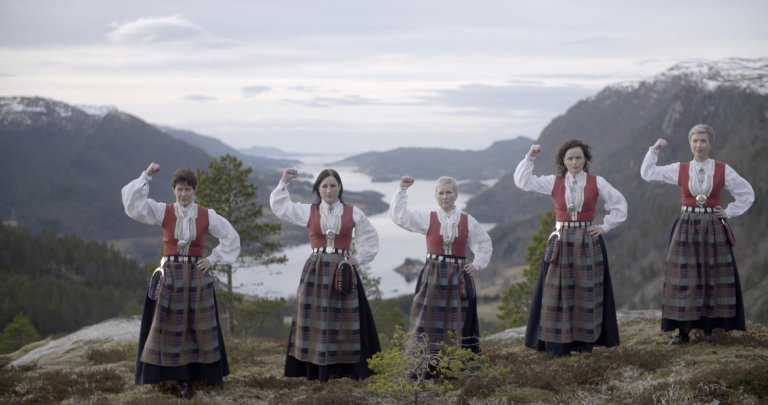 foto. fem kvinner kledt i bunad står på en fjelltopp med utsikt over en fjord og hever venstre knyttneve i været