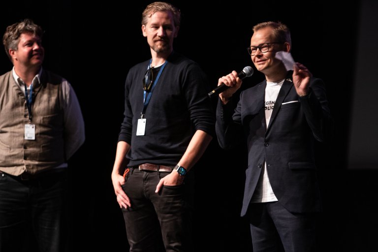 foto. tre menn i mørke klær står på scenen med mannen til høyre med mikrofon