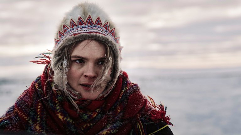 foto. ung kvinne i pelslue med samisk mønster ser ut i et snødekt landskap