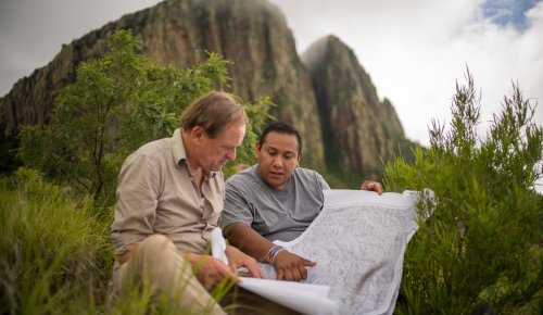 foto. to menn sitter i grønne busker foran et fjell og studerer et kart 