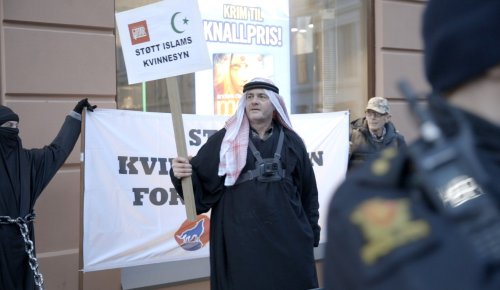 foto. mann kledd i svart kjortel og kefijeskjerf står på gata med en plakat med påskriften Støtt islams kvinnesyn, på magen har han et kamera, hver side av ham en kvinne utkledd i niqab og en mann i kamuflasjeklær. i forgrunnen en politimann 