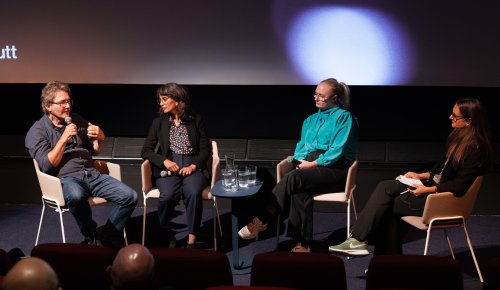 foto. fire personer sitter i hver sin stol på scene, en mann til venstre og tre kvinner, mann snakker i mikrofonen 
