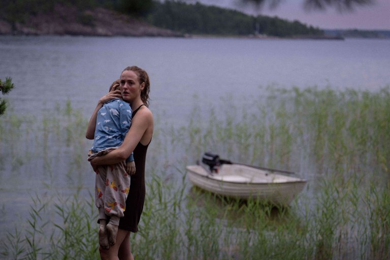 Kvinne står i sivet ved et vann og holder et lite barn inntil seg.