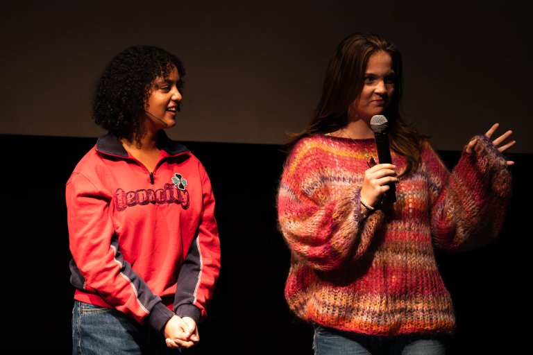 foto. ung kvinne i rød genser til venstre på bildet til høyre ung kvinne i rød strikkegenser med mikrofon på scene foran hvitt lerret