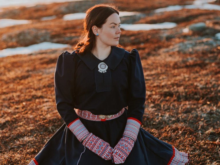 Vi ser en kvinne sitte på rødbrunt gress eller lav. Hun har en blå drakt med broderier hentet fra samisk tradisjon. Hun har også mønstrete votter og turkise øredobber. Hun har lagt brunt hår og ser til siden.