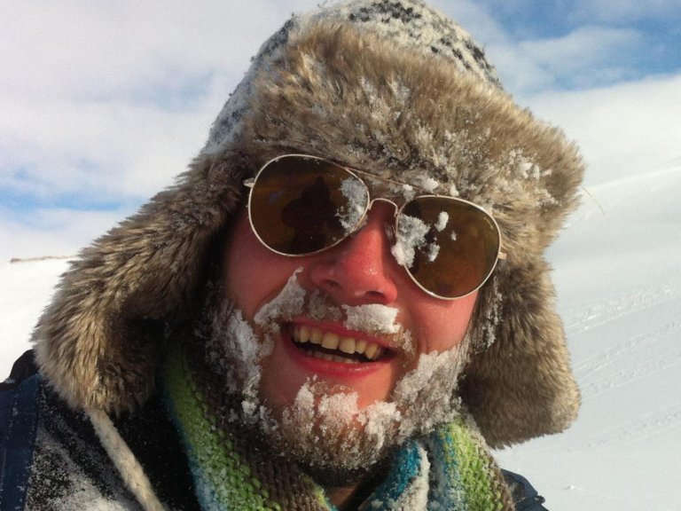 Foto: Portrett av en mann utendørs. Han har en stor lue med ørevarmere, solbriller og masse snø og is i skjegg og bart. Vi ser snølandskap i bakgrunnen.
