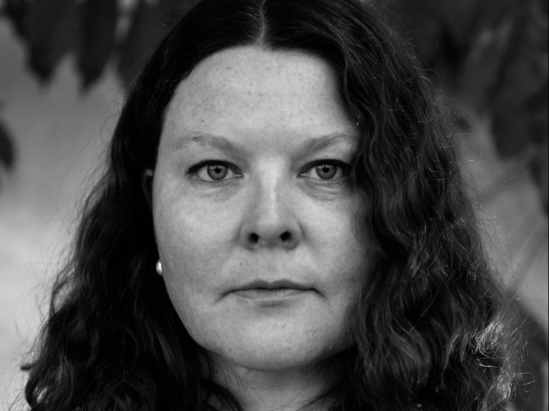 Et portrett av en kvinne fra halsen og opp. Bildet er i sort-hvitt. Hun har langt mørkt, bølgete hår i midtskill. Foto.